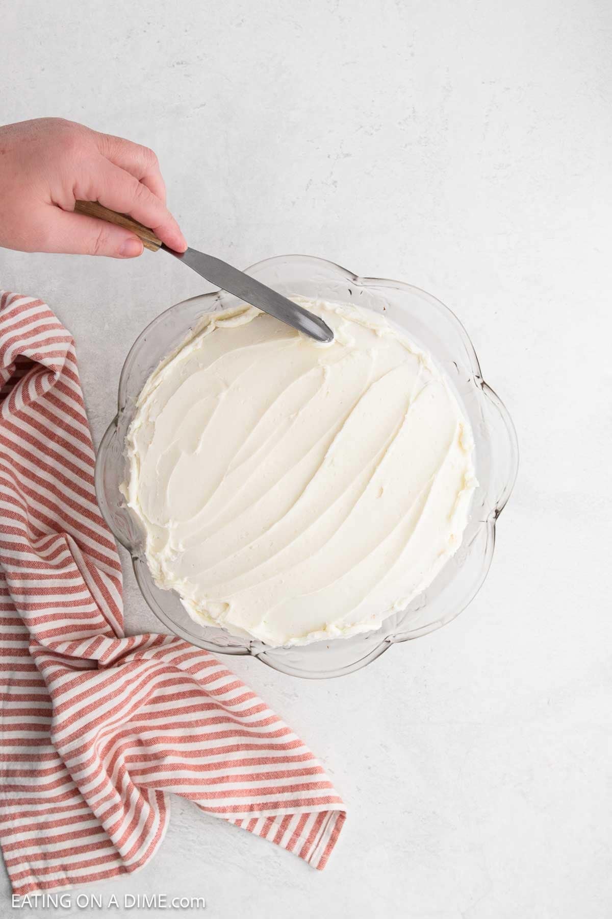 A hand uses a spatula to spread white frosting over a Strawberry Crunch Cake in a round glass dish, with a red and white striped towel beside it on a light surface.