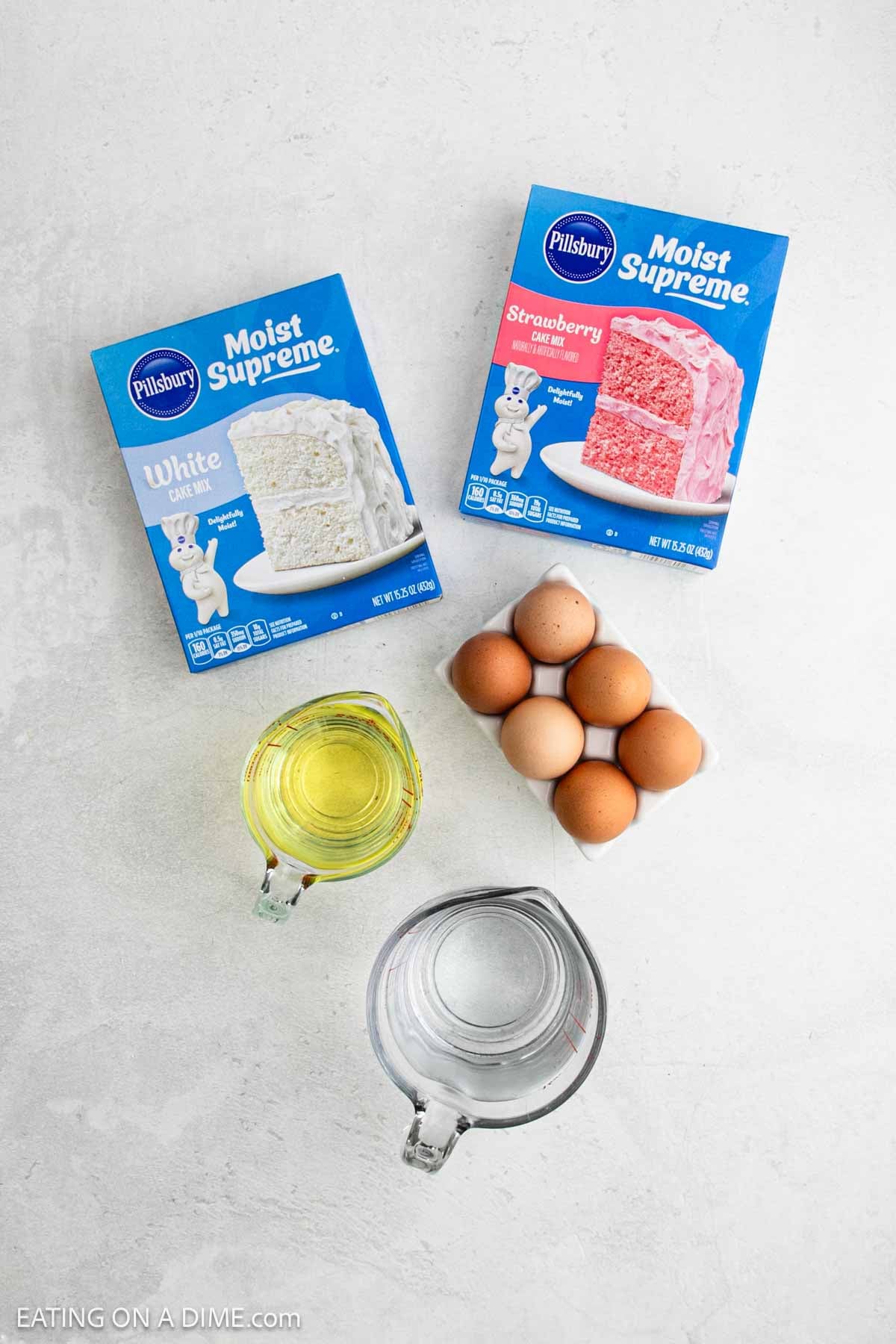 Two boxes of Pillsbury cake mix (White and Strawberry), a glass measuring cup of water, a clear container of oil, and six brown eggs—everything you need for a delicious homemade Strawberry Crunch Cake—are arranged on a light countertop.
