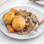 A plate with two whole potatoes, sliced carrots, and Crock Pot Chuck Roast topped with gravy and chopped parsley, with a fork on the side.