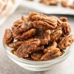 A clear glass bowl filled with Stovetop Candied Pecans sits on a gray surface, with more pecans blurred in the background. The pecans appear coated in a light, crunchy sugar glaze.