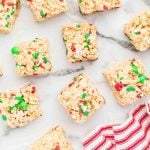 Squares of Christmas Rice Krispie Treats, topped with red and green M&M candies and colorful sprinkles, are arranged on a white marble surface. A red and white striped cloth peeks in from the corner.