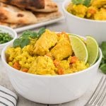 A white bowl filled with Crock Pot Chicken Curry Rice Bowls—yellow rice, chicken pieces, and diced carrots, garnished with cilantro and two lime wedges. In the background are naan bread pieces and another bowl of the dish.