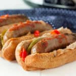 Three Crockpot Sausage and Peppers sandwiches in whole wheat buns, topped with sautéed onions, bell peppers, and tomato sauce, are arranged in a row on a white surface with a blue patterned cloth in the background.