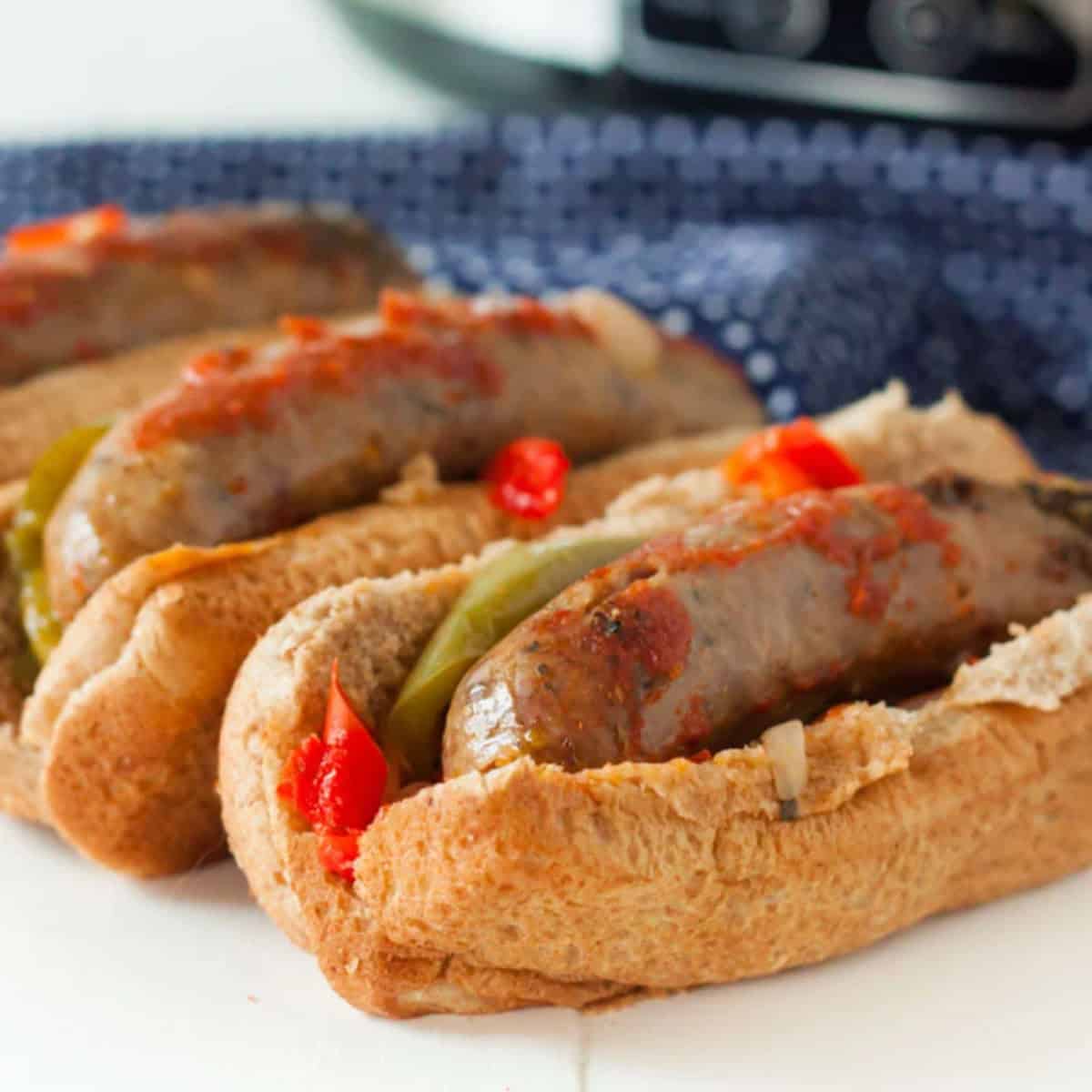 Three Crockpot Sausage and Peppers sandwiches in whole wheat buns, topped with sautéed onions, bell peppers, and tomato sauce, are arranged in a row on a white surface with a blue patterned cloth in the background.
