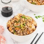 A white bowl filled with Egg Roll in a Bowl—a mixture of ground meat, shredded cabbage, and carrots—sits on a marble surface near black chopsticks, a jar of soy sauce, another bowl, and sliced green onions.