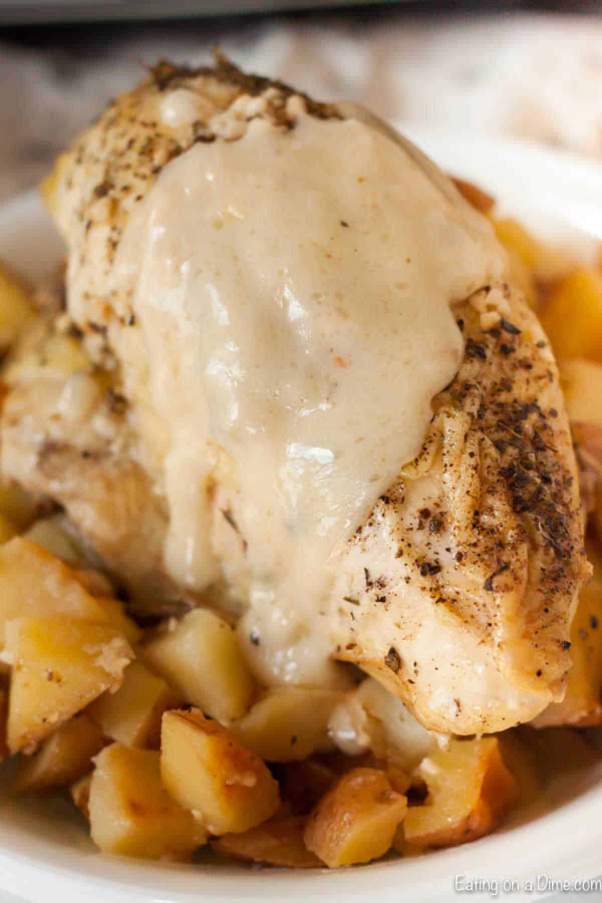 A close-up of a seasoned chicken breast topped with creamy gravy, reminiscent of a slow cooker lemon garlic chicken recipe, served on a bed of diced, roasted potatoes. Browned potatoes add texture and contrast to the dish's light golden hues.