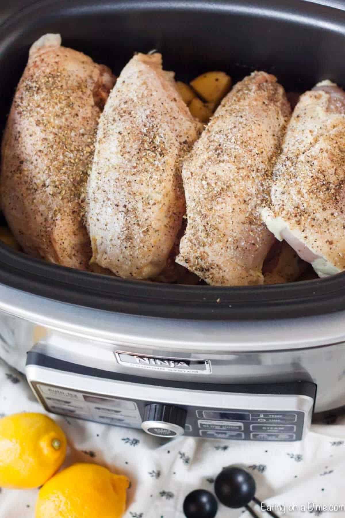 Chicken breasts seasoned with spices create a delightful aroma in the slow cooker. Nearby, bright lemons and black olives sit on a patterned cloth, promising a delicious lemon garlic chicken recipe that’s both simple and savory.