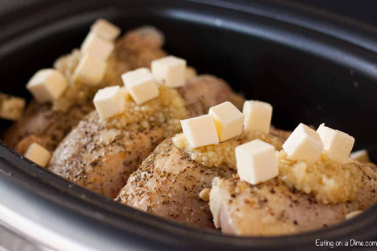 Raw seasoned chicken breasts in a slow cooker, topped with minced garlic, cubes of butter, and a hint of lemon for an exquisite lemon garlic chicken recipe.
