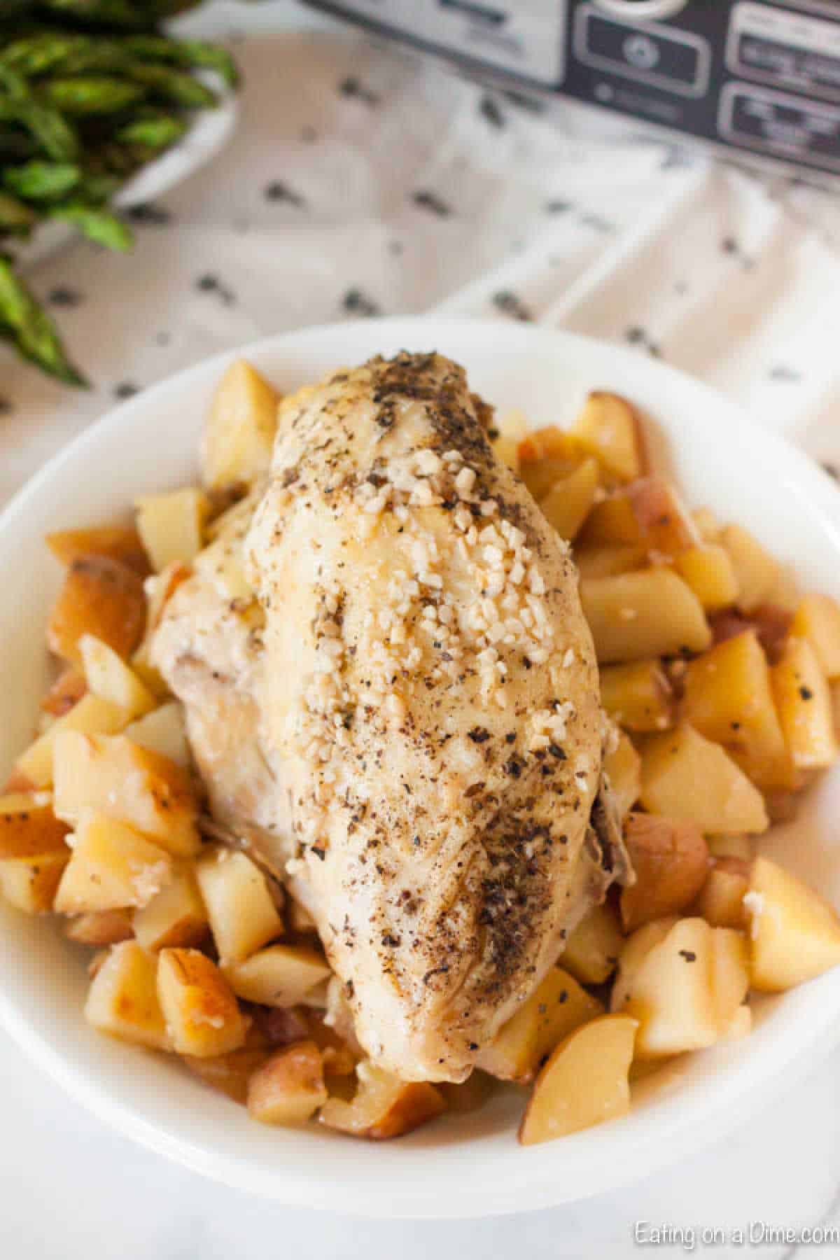 A plate showcasing a slow cooker lemon garlic chicken breast seasoned with herbs, nestled atop diced potatoes. The dish is placed near a cloth napkin with a hint of greens in the background, creating an inviting meal setting.