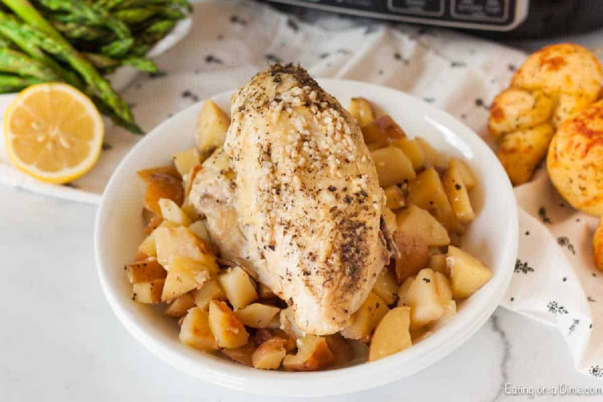 A white plate showcases a seasoned chicken breast on a bed of diced potatoes, reminiscent of a slow cooker lemon garlic chicken recipe. Roasted asparagus and a halved lemon enhance the scene, with two bread rolls in the background. The dish is beautifully garnished with herbs.