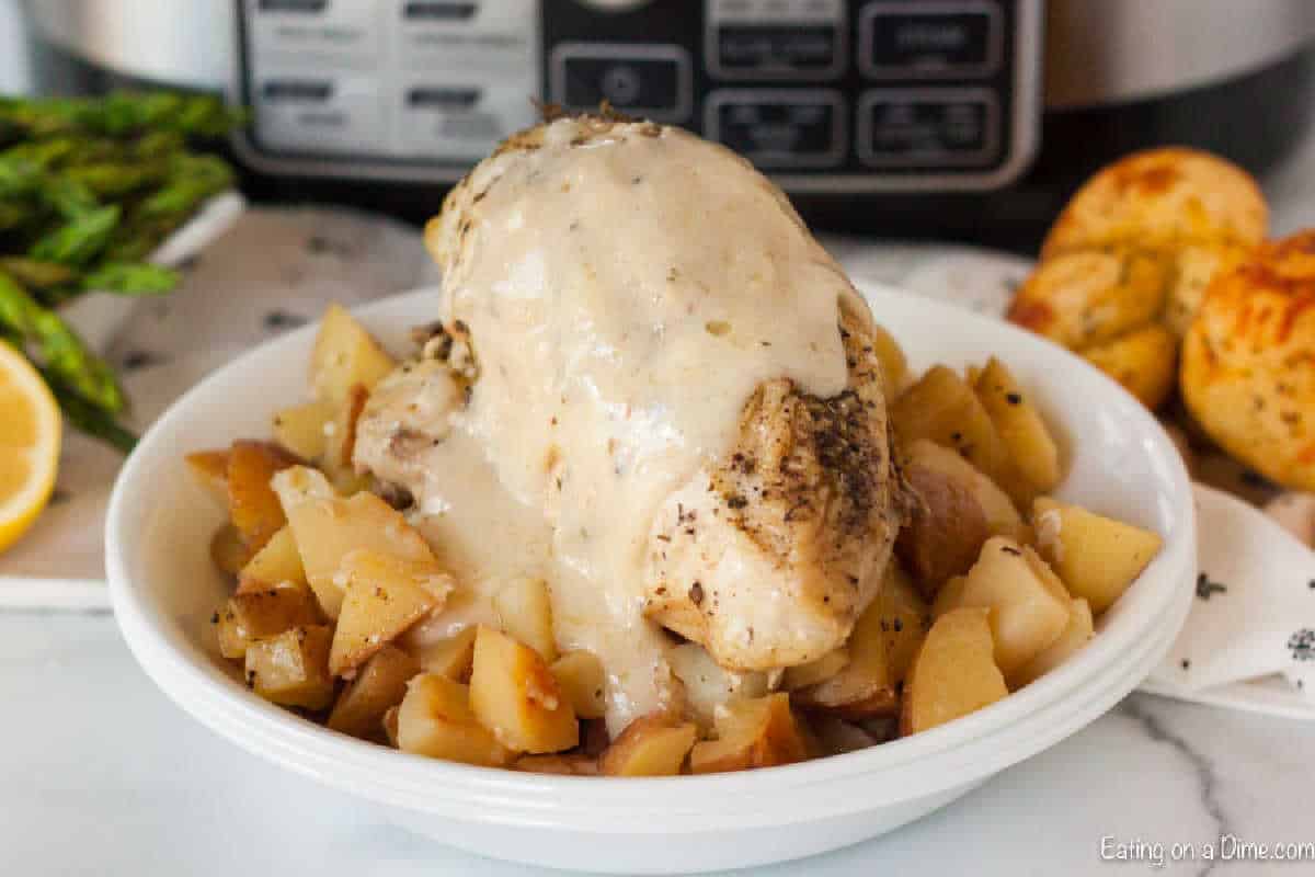 A bowl of creamy Italian slow cooker lemon garlic chicken with diced potatoes is topped with sauce. On the side, asparagus spears, a lemon slice, and a bread roll rest on a marble surface.
