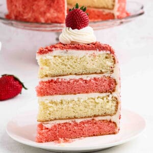 A slice of layered Strawberry Crunch Cake with strawberry and vanilla, covered in white frosting, topped with a swirl of cream and a whole strawberry, sits on a white plate. The moist cake is decorated with pink crumbs.