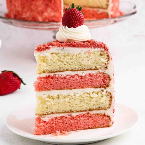 A slice of layered Strawberry Crunch Cake with strawberry and vanilla, covered in white frosting, topped with a swirl of cream and a whole strawberry, sits on a white plate. The moist cake is decorated with pink crumbs.