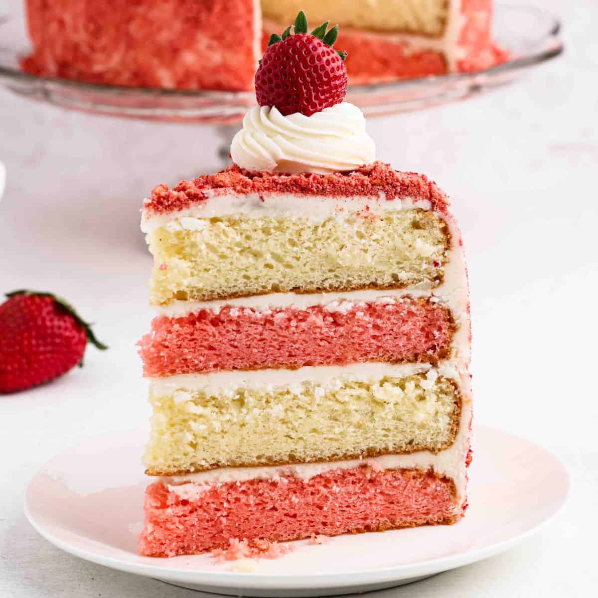 A slice of layered Strawberry Crunch Cake with strawberry and vanilla, covered in white frosting, topped with a swirl of cream and a whole strawberry, sits on a white plate. The moist cake is decorated with pink crumbs.