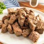 A white plate filled with savory Air Fryer Steak Bites—cooked, bite-sized steak pieces seasoned with herbs and minced garlic, with a striped blue napkin and parsley blurred in the background.