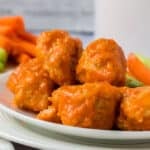 A plate of saucy buffalo chicken meatballs, inspired by boneless chicken wings, is served with celery and carrot sticks in the background.