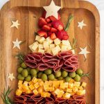 A wooden christmas tree charcuterie board features food arranged in a festive tree shape: cheese stars on top, layers of strawberries, cheese cubes, folded salami, green olives, cheddar cubes, pepperoni, plus rosemary and extra cheese stars for decoration.