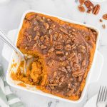 A square white baking dish filled with Cracker Barrel Sweet Potato Casserole Recipe topped with pecans and cinnamon. A serving has been scooped out, revealing the creamy orange filling, with pecans scattered on the marble surface nearby.