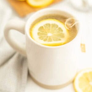 A white mug filled with tea, reminiscent of a Starbucks Medicine Ball Recipe, is topped with a floating lemon slice and a tea bag string hanging over the side, sitting on a light-colored cloth with a lemon slice nearby.