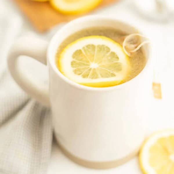 A white mug filled with tea, reminiscent of a Starbucks Medicine Ball Recipe, is topped with a floating lemon slice and a tea bag string hanging over the side, sitting on a light-colored cloth with a lemon slice nearby.