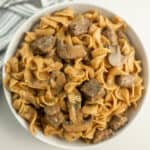 A white bowl brimming with Instant Pot beef stroganoff showcases egg noodles, sliced mushrooms, and tender beef, all enveloped in a creamy sauce. Garnished with chopped herbs, the dish rests on a white surface with a striped cloth peeking into view.