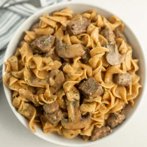 A white bowl brimming with Instant Pot beef stroganoff showcases egg noodles, sliced mushrooms, and tender beef, all enveloped in a creamy sauce. Garnished with chopped herbs, the dish rests on a white surface with a striped cloth peeking into view.