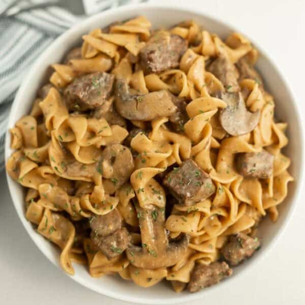 A white bowl brimming with Instant Pot beef stroganoff showcases egg noodles, sliced mushrooms, and tender beef, all enveloped in a creamy sauce. Garnished with chopped herbs, the dish rests on a white surface with a striped cloth peeking into view.