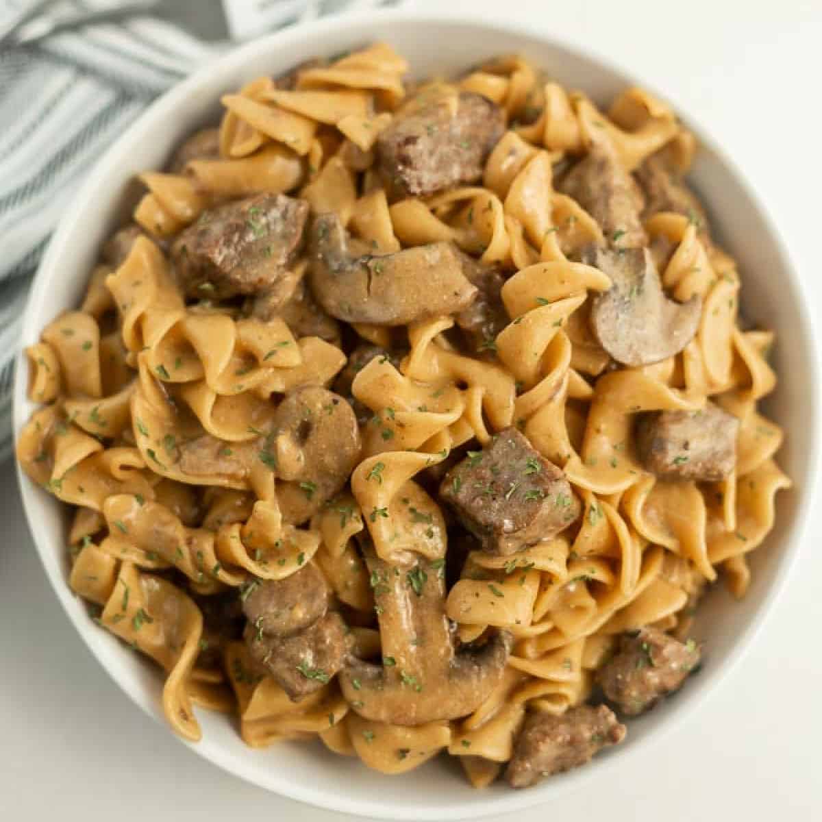 A white bowl brimming with Instant Pot beef stroganoff showcases egg noodles, sliced mushrooms, and tender beef, all enveloped in a creamy sauce. Garnished with chopped herbs, the dish rests on a white surface with a striped cloth peeking into view.