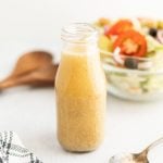 A glass bottle filled with light brown Olive Garden Salad Dressing sits on a white surface. In the background, there is a clear bowl of salad with tomatoes, olives, and lettuce, as well as a wooden spoon.