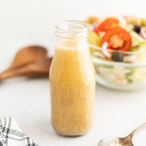 A glass bottle filled with light brown Olive Garden Salad Dressing sits on a white surface.
