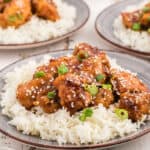 Crispy sesame chicken over white rice on a plate topped with green onions and sesame seeds.