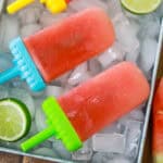 Two homemade watermelon popsicles with green and blue handles rest on ice cubes in a metal tray, surrounded by slices of fresh lime.