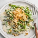 A plate of green bean and asparagus casserole topped with crispy breadcrumbs and grated cheese, covered in creamy white sauce, with a fork on the side.
