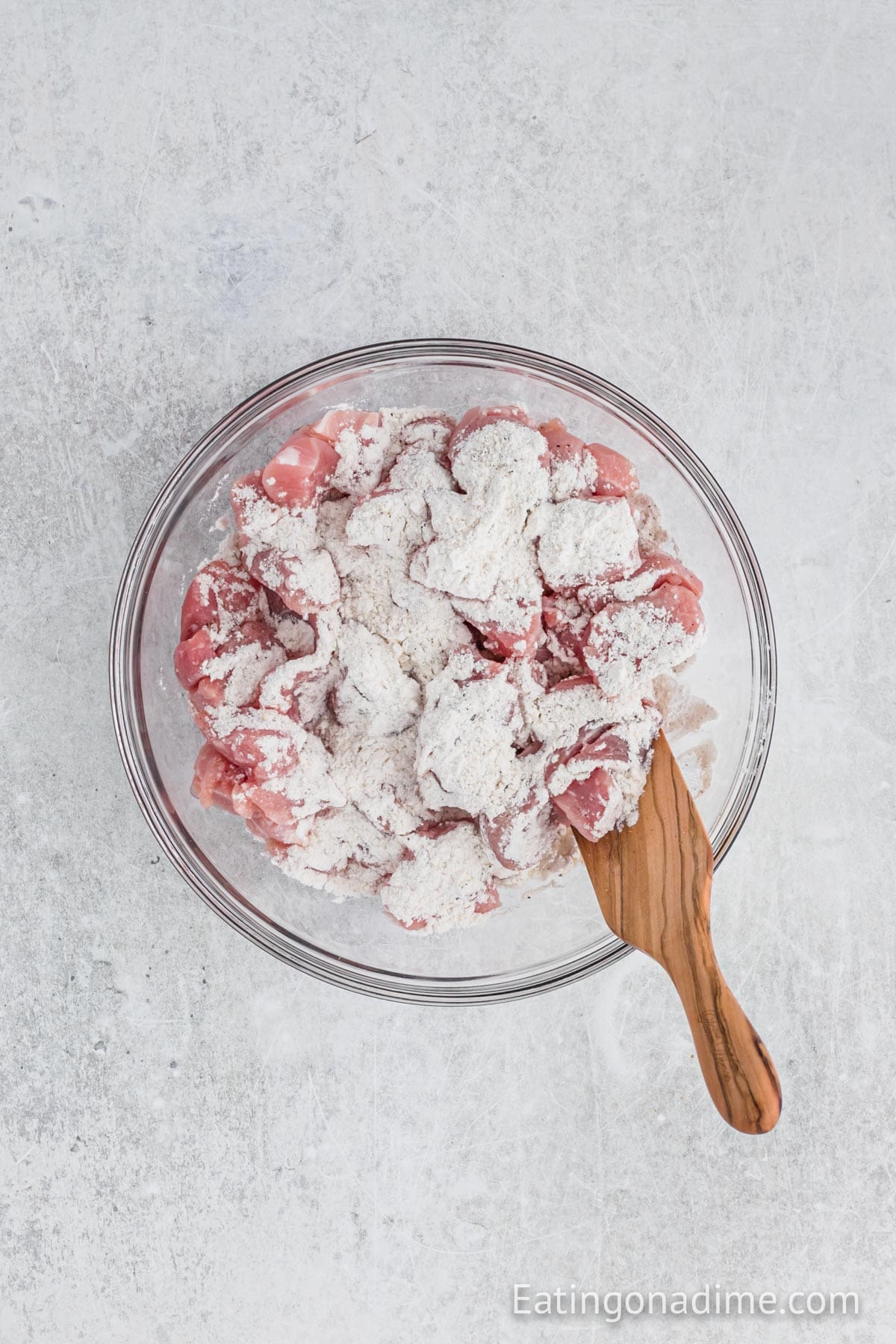 A glass bowl filled with raw, cubed meat coated in flour sits on a light gray surface, ready to become a hearty Slow Cooker Pork Stew. A wooden spatula rests in the bowl, prepared for the next step.