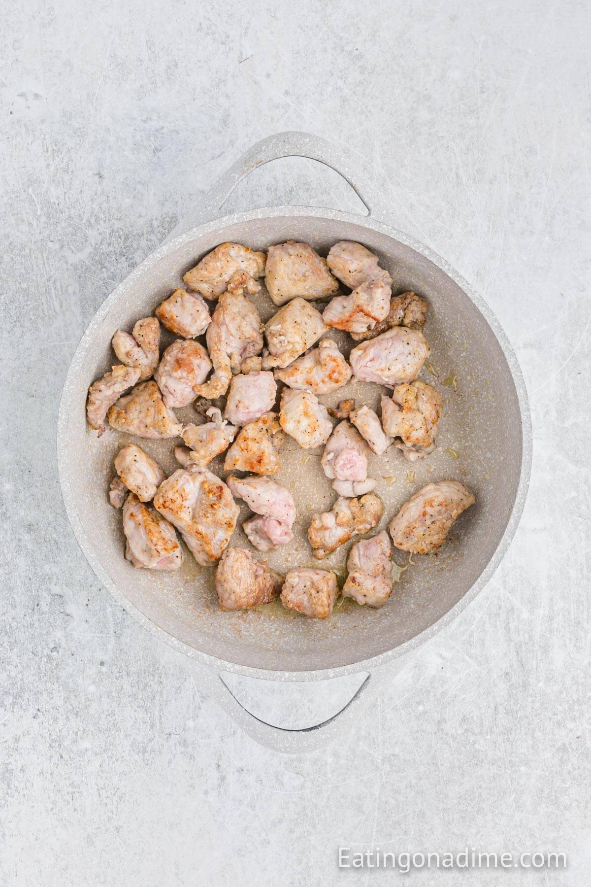 A white pot filled with browned chunks of meat, perfect for making Slow Cooker Pork Stew, sits on a light gray textured surface. The pot handles are visible, and a watermark reads "Eatingonadime.com" in the bottom right corner.