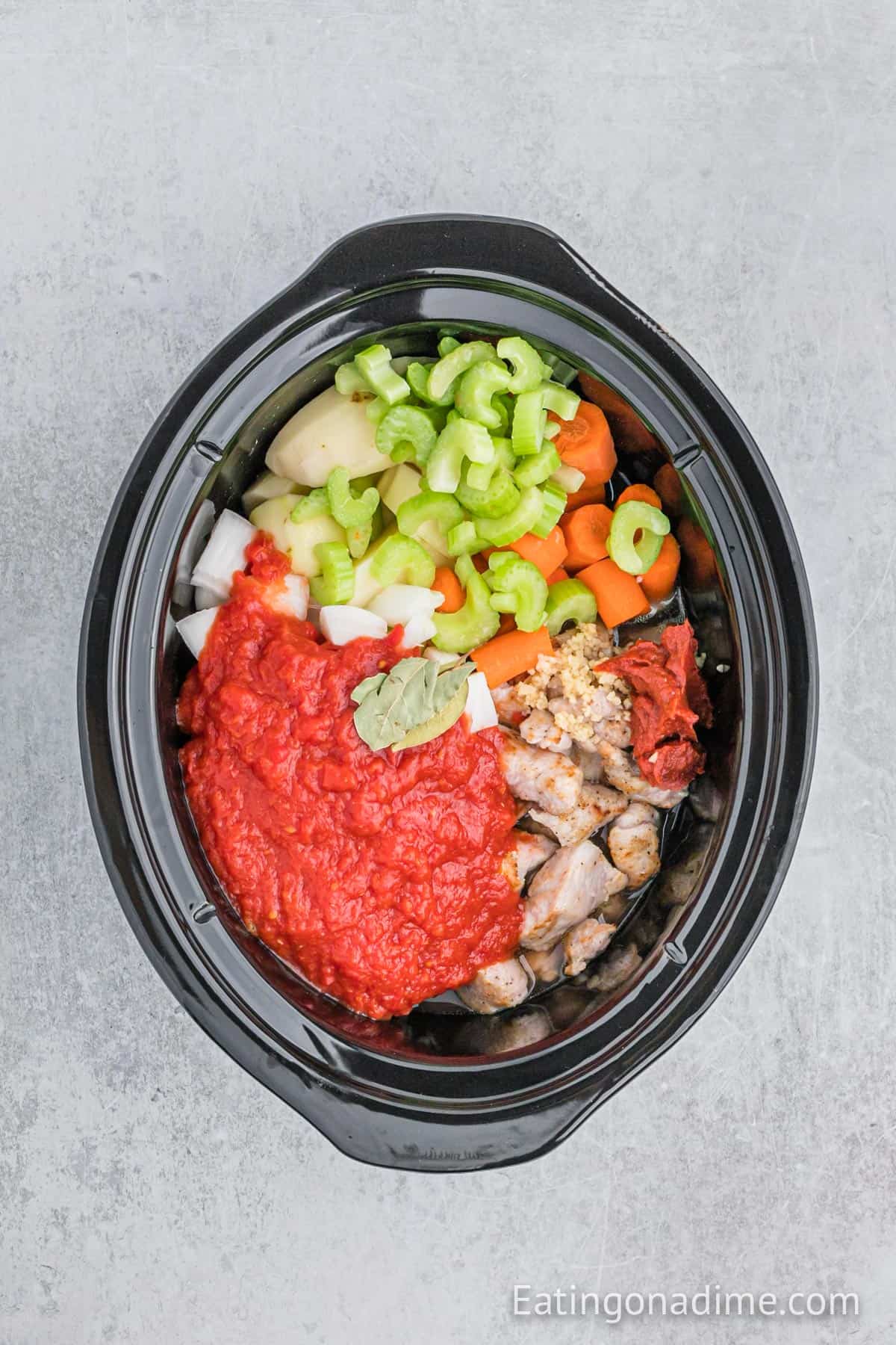 A slow cooker filled with ingredients for a hearty Slow Cooker Pork Stew: chunks of raw chicken, sliced carrots, celery, onions, potatoes, all topped with crushed tomatoes, tomato paste, and a bay leaf—ready to be cooked.