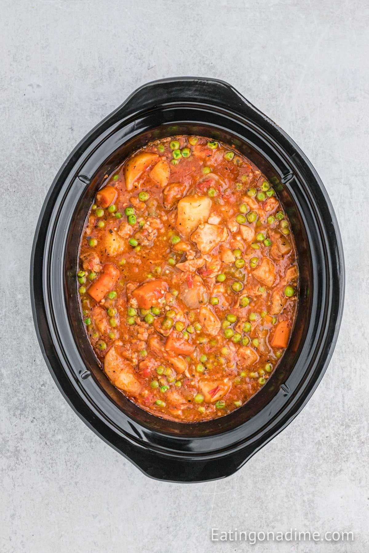 Overhead view of a slow cooker filled with Slow Cooker Pork Stew, featuring tender pork, potatoes, carrots, peas, and a tomato-based broth on a light gray background.