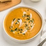A bowl of creamy Crock Pot Sweet Potato Soup, garnished with a swirl of cream, pumpkin seeds, and chopped herbs, sits on a white surface next to a beige napkin and a wooden cutting board.