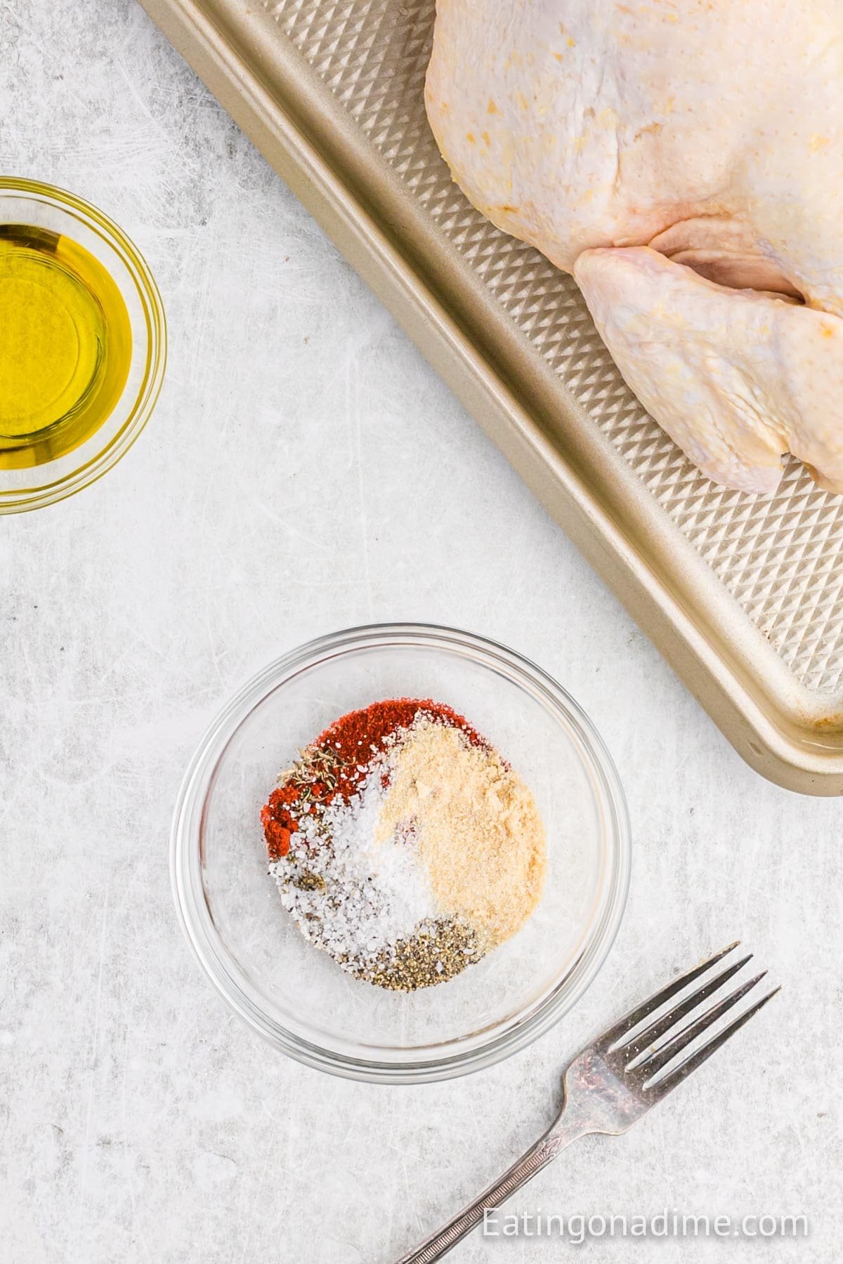 Glass bowl with assorted spices, a fork, a bowl of olive oil, and raw Rotisserie Chicken on a baking tray displayed on a light countertop.
