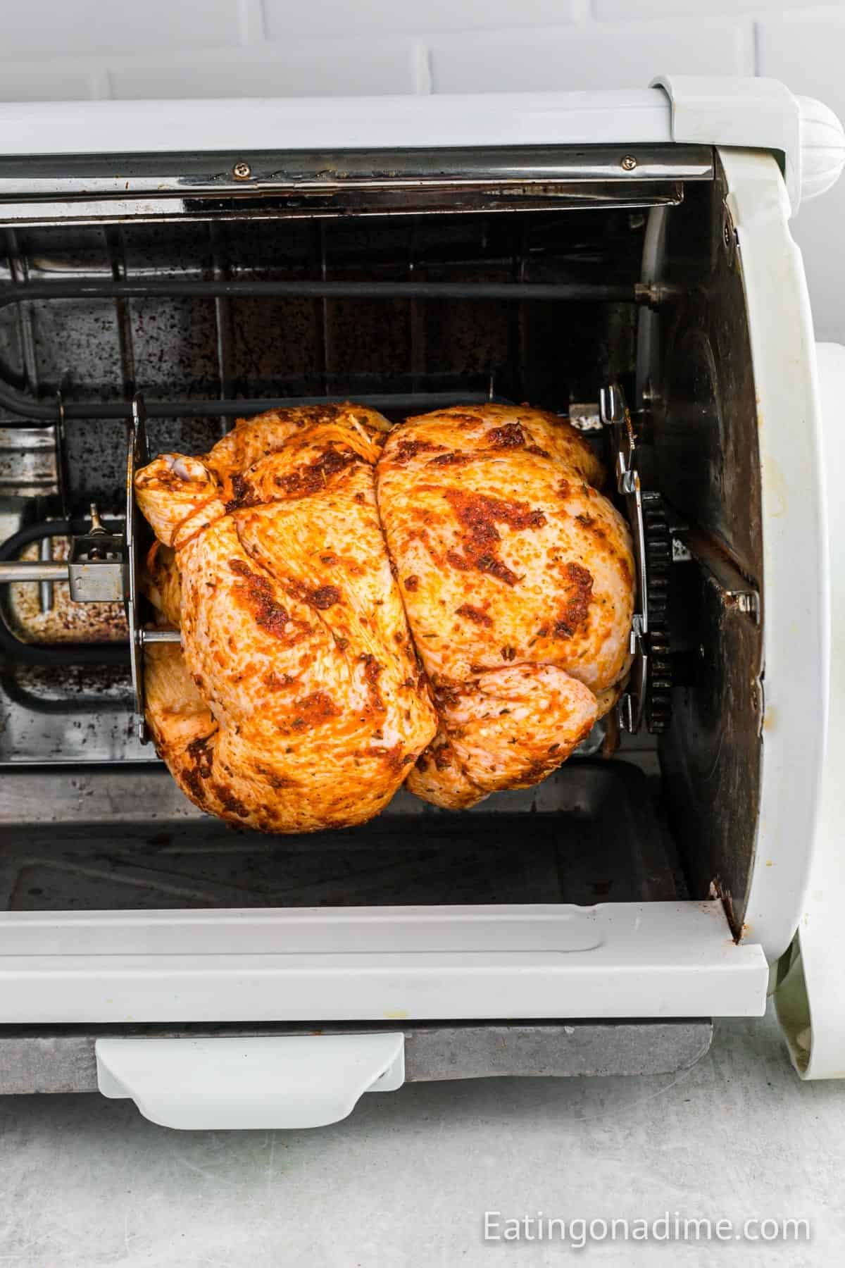 A seasoned Rotisserie Chicken is roasting on a spit inside an electric rotisserie oven, with the brand name "Eatingonadime.com" visible in the corner.