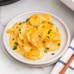 A plate of creamy, cheesy Instant Pot Scalloped Potatoes garnished with chopped fresh herbs, served on a white plate with a striped napkin beside it.