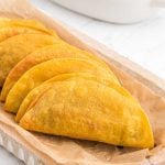 Five crispy, golden-brown Taco Dorados are lined up on a piece of parchment paper on a wooden tray, their folded shells showing a slight texture and light brown spots.
