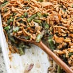 A close-up of a Green Bean Casserole with Bacon Recipe, topped with crispy fried onions and partially served with a wooden spoon in a white baking dish. The creamy filling, green beans, and bits of bacon peek out from under the crunchy topping.