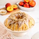 A Peach Cobbler Pound Cake with white glaze and crumb topping sits on a cake stand. Fresh peaches and a dish of chopped nuts are in the background on a white countertop.