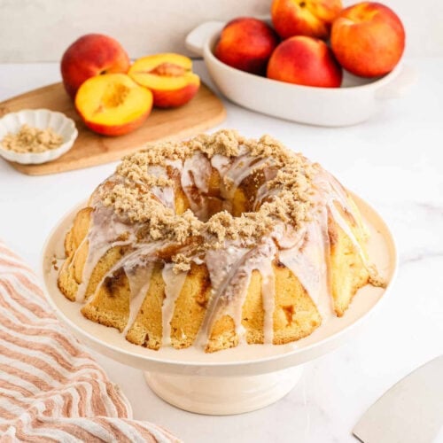 A Peach Cobbler Pound Cake with white glaze and crumb topping sits on a cake stand. Fresh peaches and a dish of chopped nuts are in the background on a white countertop.