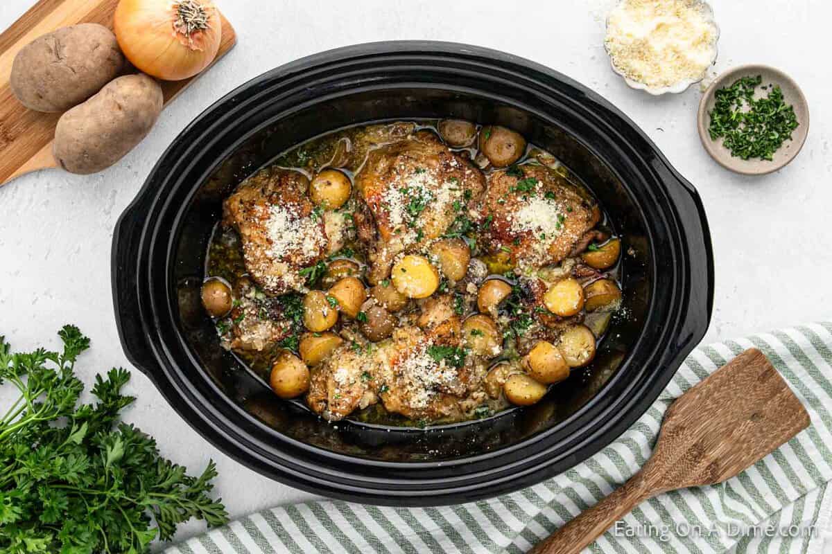 A black slow cooker filled with Slow Cooker Garlic Parmesan Chicken: tender chicken thighs, baby potatoes, and a flavorful broth, garnished with fresh parsley and grated Parmesan cheese. Surrounding the cooker are fresh parsley, a striped kitchen towel, an onion, potatoes, and a small dish of Parmesan.