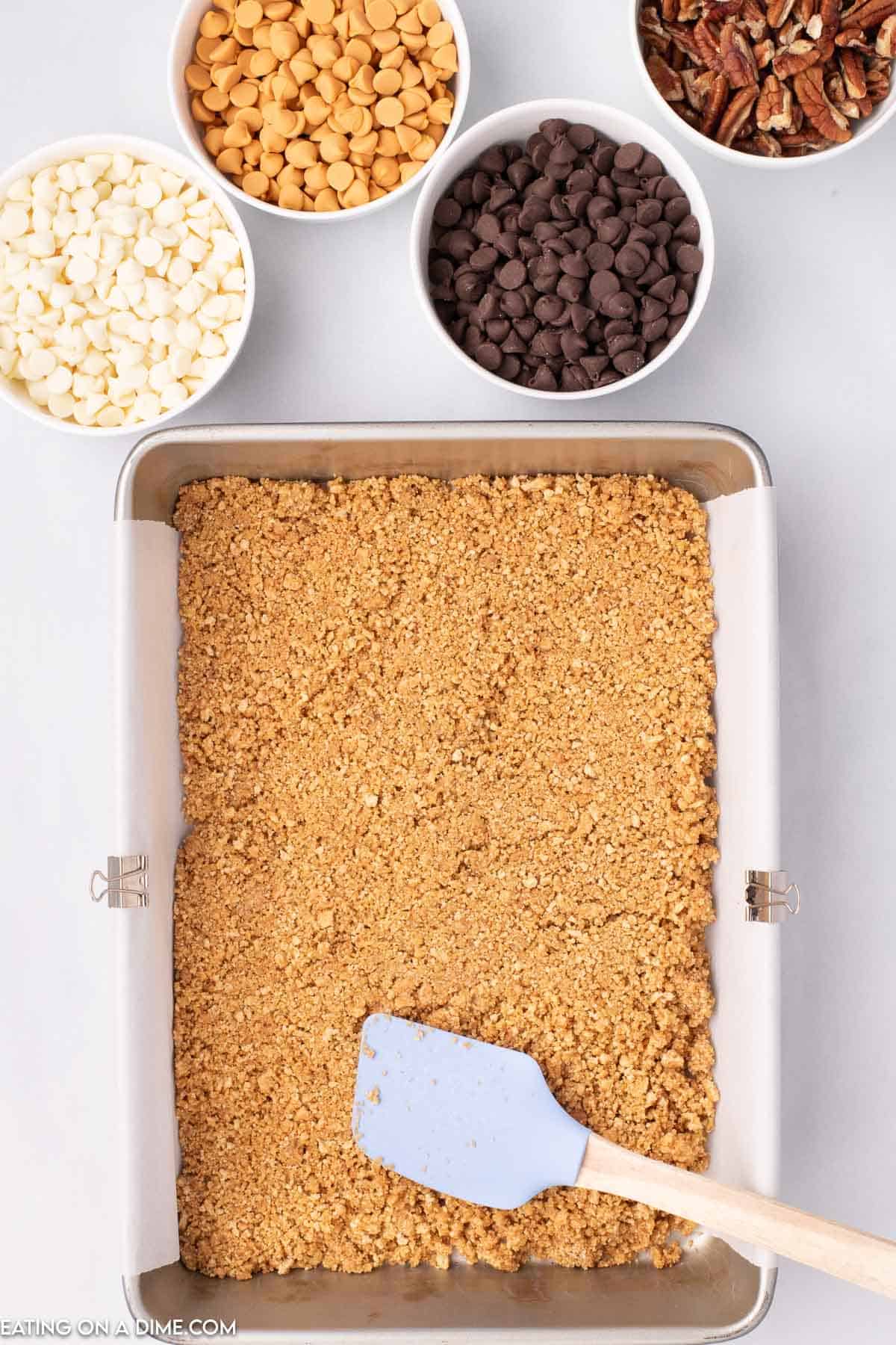 Pressing the graham cracker crumbs in a baking dish with a spatula and bowl of chocolate chips and pecans on the side