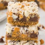 A close-up of three stacked Seven Layer Bars with a crumbly base, layers of chocolate chips, butterscotch chips, nuts, and shredded coconut. The bars look chewy and rich, with visible textures and a golden brown crust.