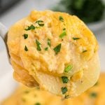 Close-up of creamy Au Gratin Potatoes Crock Pot Recipe, topped with melted cheese and garnished with chopped fresh parsley, held up on a serving utensil.