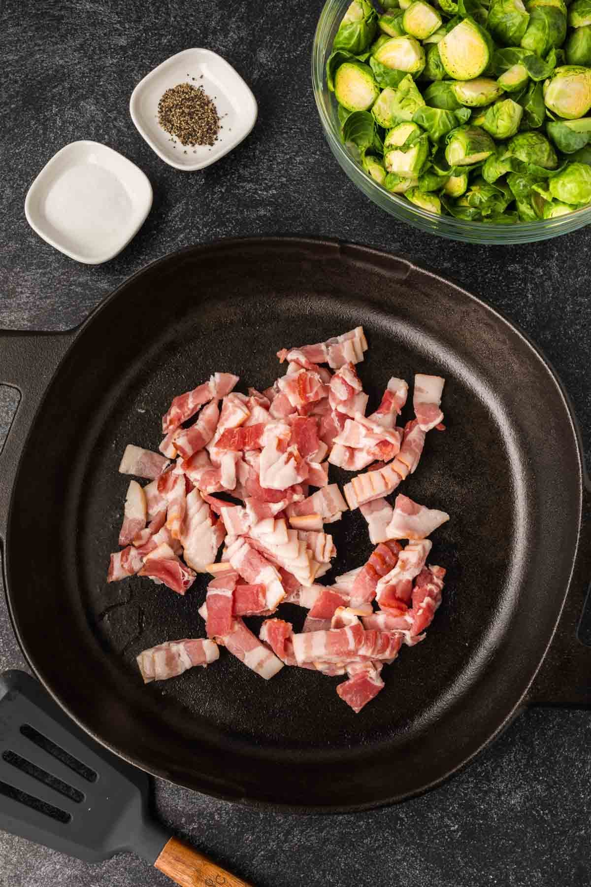 A cast iron skillet with chopped raw bacon pieces, surrounded by a bowl of halved Brussels sprouts ready for Bacon Brussel Sprouts, plus a spatula and small dishes of salt and pepper on a dark countertop.